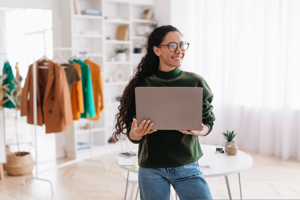 cheerful female clothing designer holding laptop working online in studio